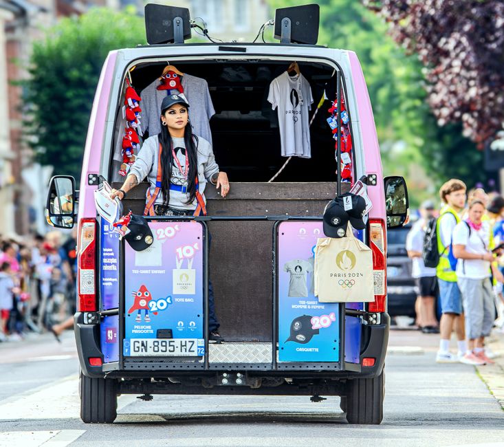 Mulher vendendo produtos relacionados às Olimpíadas de Paris 2024 em uma van de rua durante evento ao ar livre.