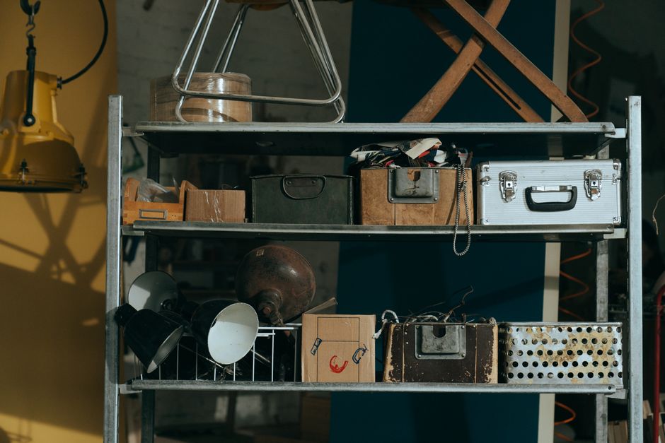 Imagem de uma prateleira de depósito com objetos variados, incluindo caixas, ferramentas e utensílios antigos, sugerindo um ambiente de armazenamento ou oficina.
