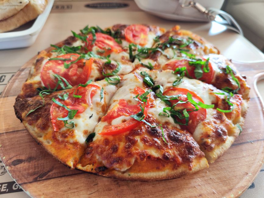 Pizza de marguerita com molho de tomate, queijo derretido e folhas de manjericão, servida sobre uma tábua de madeira em um ambiente informal.
