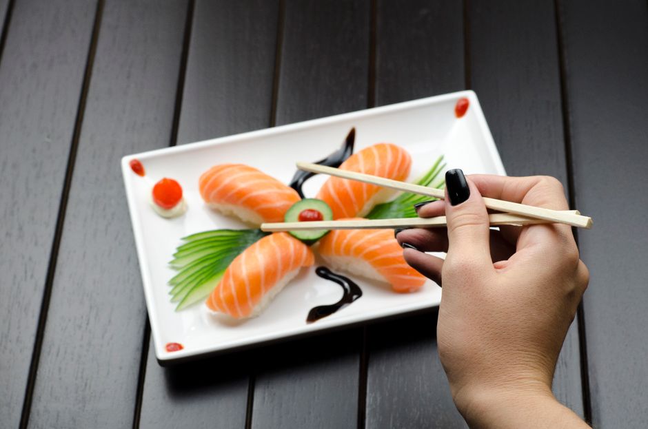 Pessoa usando hashis para pegar sushi em um prato com sushi, wasabi e gengibre, destacando a gastronomia japonesa.