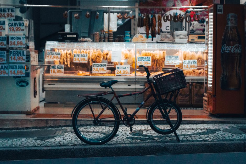 Imagem de uma bicicleta estacionada na calçada em frente a uma lanchonete que vende petiscos e snacks, capturando a atmosfera urbana noturna.