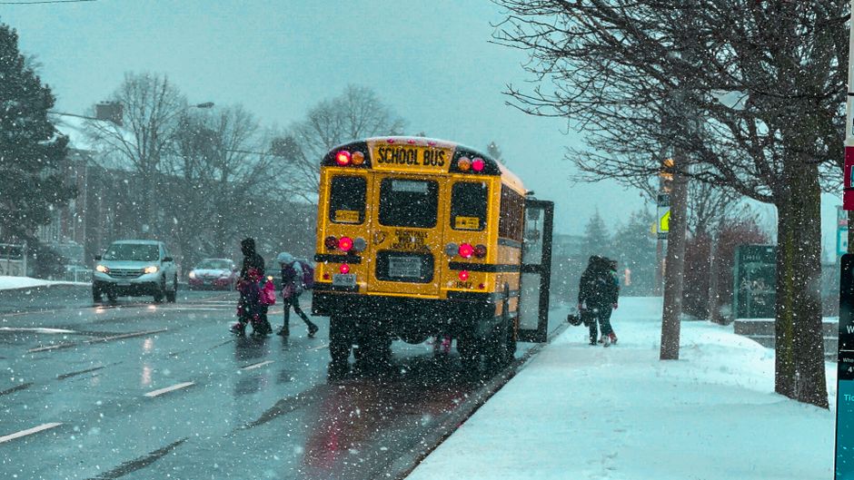 Cena de escola com estudantes e transporte escolar durante uma nevasca na rua, destacando o clima de inverno e a rotina escolar.