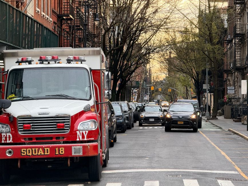 Uma rua urbana com carros estacionados de ambos os lados e uma viatura de bombeiros na frente, com árvores e edifícios ao fundo ao entardecer.