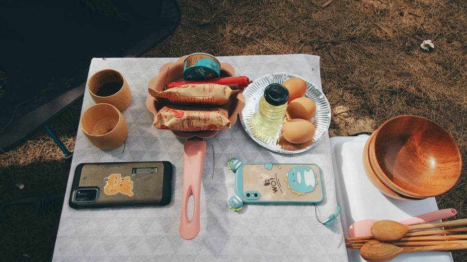 Mesa ao ar livre com café, ovos, utensílios de cozinha, celulares e itens para refeições, sugerindo um momento de descanso ou lazer ao ar livre.