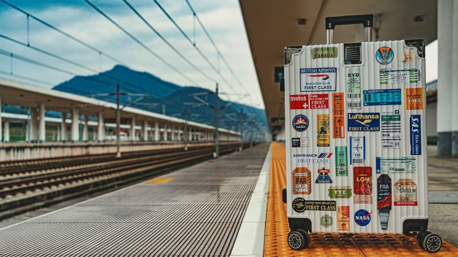 Mala de viagem decorada com adesivos de companhias aéreas e destinos turísticos, pronta para uma jornada no aeroporto ao lado de trilhos e montanhas ao fundo.