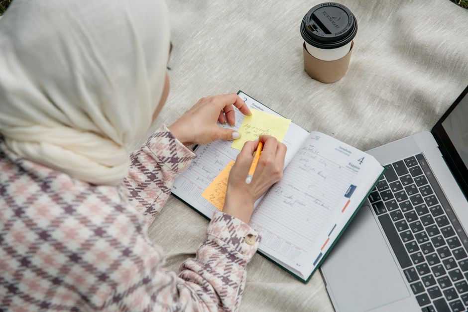 Mulher usando hijab organizando tarefas e anotando ideias em um caderno próximo ao laptop, em um ambiente de trabalho ou estudo.