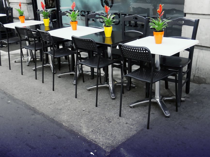 Mesas externas de café ou restaurante decoradas com vasos de flores coloridos, criando um ambiente acolhedor.
