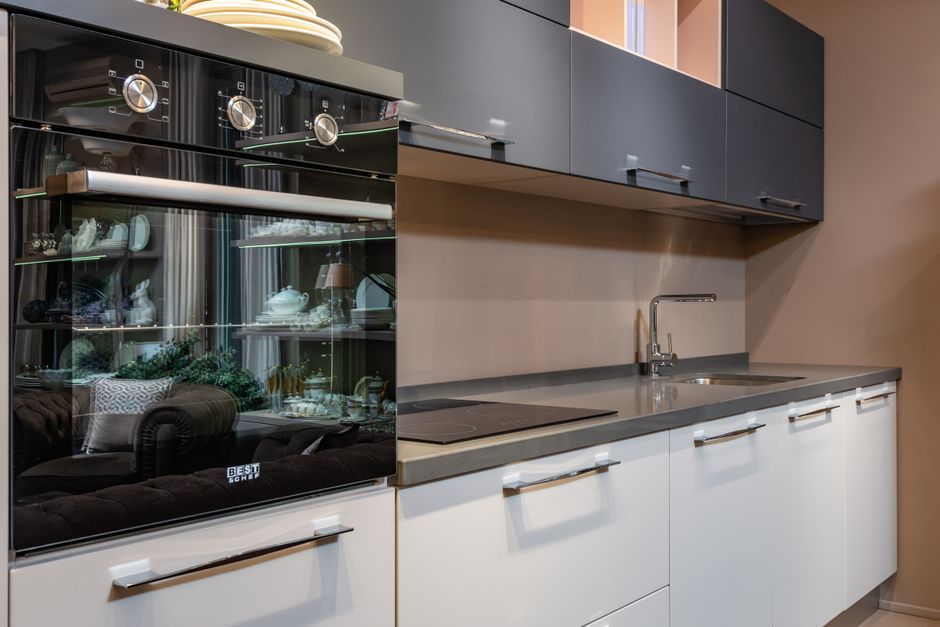 Imagem de uma cozinha moderna com armários cinza e brancos, bancada de granito e uma cristaleira com louças expostas, refletida na porta de um forno embutido.