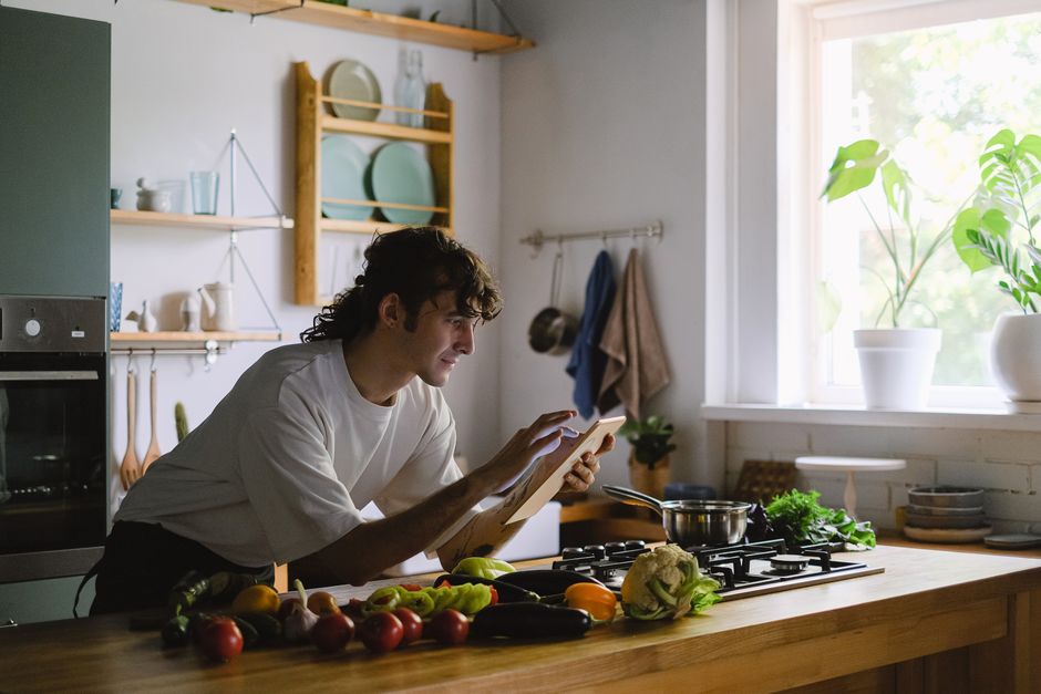 Homem usando um tablet na cozinha, rodeado por ingredientes frescos, em um ambiente bem iluminado e decorado com plantas.