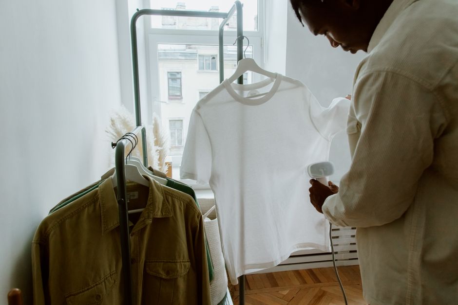 Pessoa passando roupa branca a vapor em ambiente com roupas penduradas próximo à janela durante o dia.
