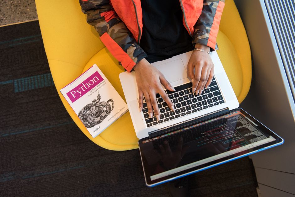 Pessoa trabalhando com programação em um laptop enquanto lê um livro de Python, em um ambiente de trabalho informal.