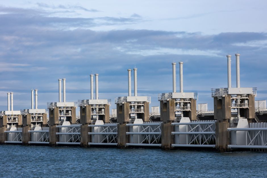 Visão de uma usina hidrelétrica com turbinas de instalação industrial ao lado de uma ampla área de água, sob céu parcialmente nublado, representando energia renovável e sustentabilidade.