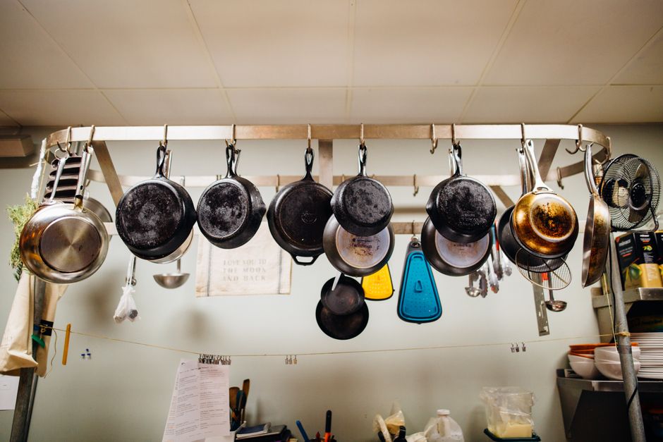 Imagem de panelas penduradas em uma cabide de cozinha, com utensílios diversos ao fundo, em ambiente doméstico ou comercial de gastronomia.