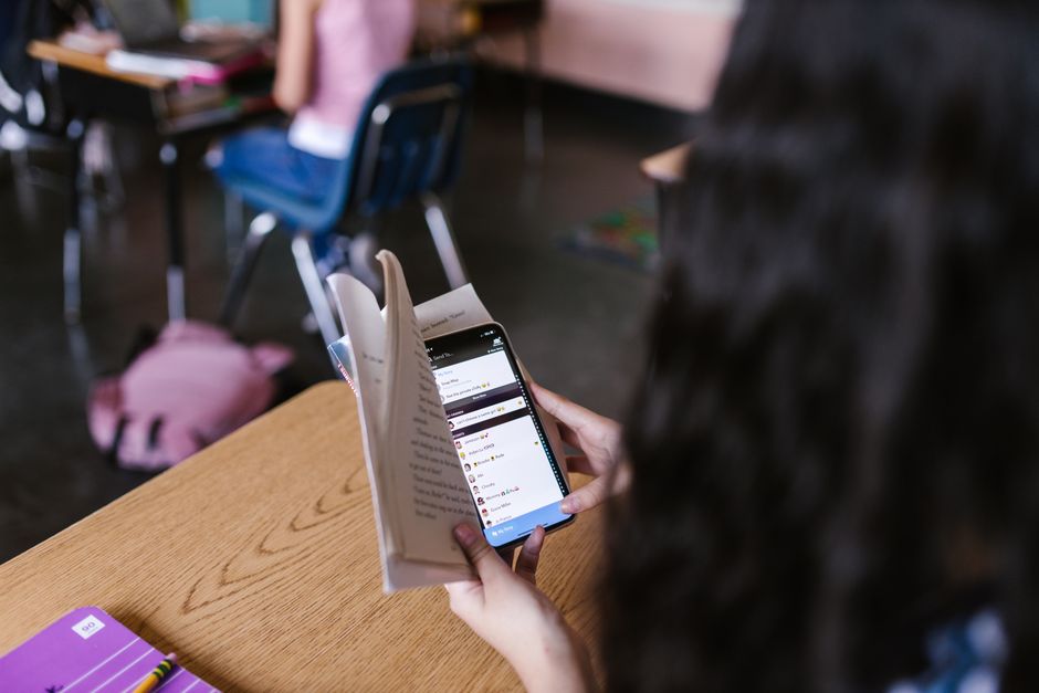 Pessoa usando smartphone na sala de aula com estudantes ao fundo, com foco na tecnologia e aprendizagem.