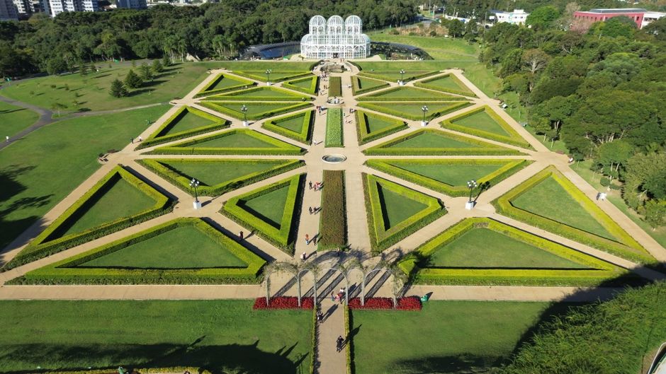 Imagem aérea de um jardim simétrico com caminhos cruzados, um pavilhão de vidro ao fundo e áreas verdes bem cuidadas, em um espaço de lazer ou parque público.