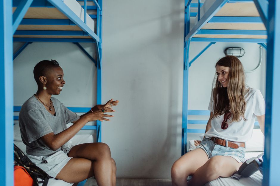 Duas jovens conversando e sorrindo dentro de um dormitório com beliches azuis, em um ambiente de convivência entre amigas.