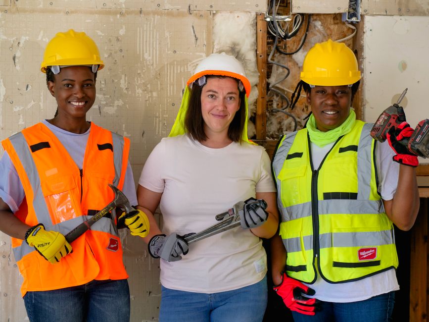 Três mulheres engenheiras ou trabalhadores da construção civil posando com sorriso, usando capacetes e vestimentas de segurança em um ambiente de construção.