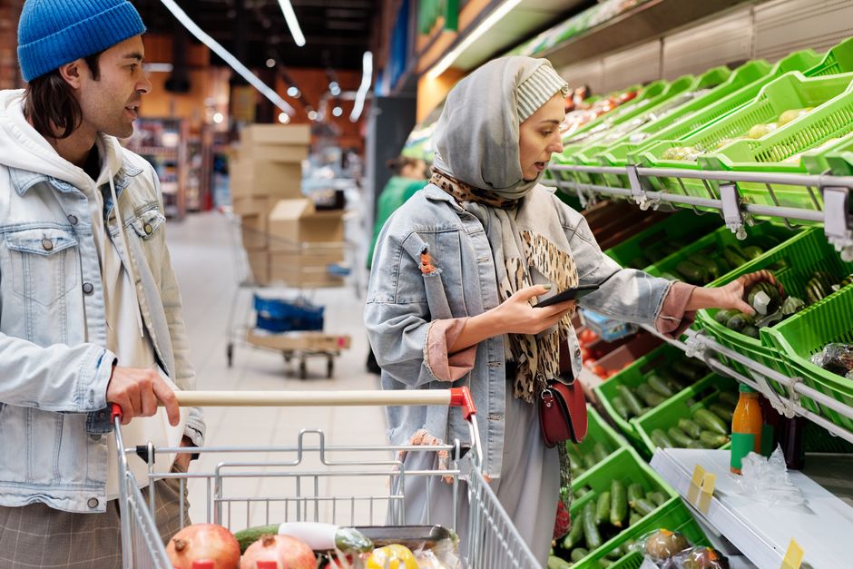 Pessoas fazendo compras de alimentos em uma feira ou supermercado, com foco na seleção de hortifrúti e na interação com produtos frescos.