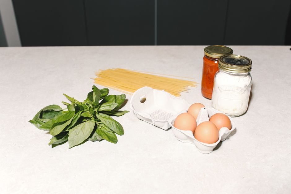 Imagem de ingredientes de cozinha como ovos, tomate, macarrão, folhas de manjericão e temperos em potes, focando no preparo de uma receita italiana.