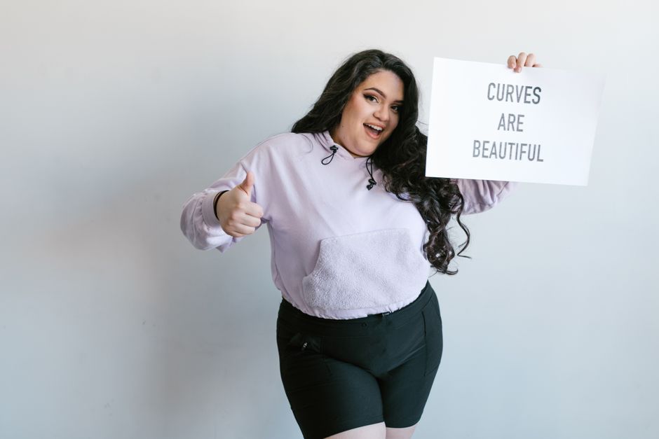 Mulher sorridente segurando um cartaz com a frase 'Curves are beautiful', transmitindo positividade corporal e autoestima.