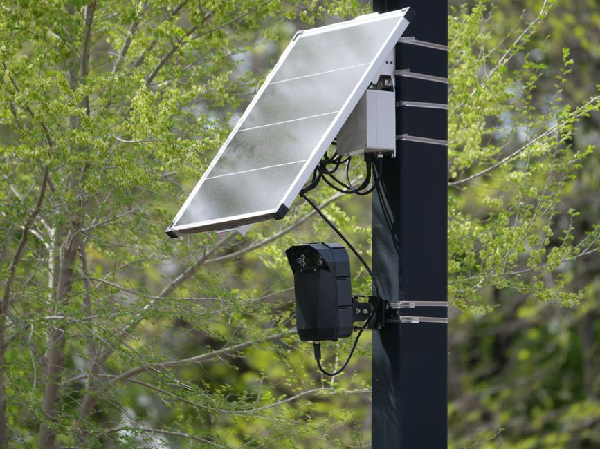 Painel solar e câmera de monitoramento instalados em um poste numa área próxima à natureza para vigilância ou estudos ambientais.