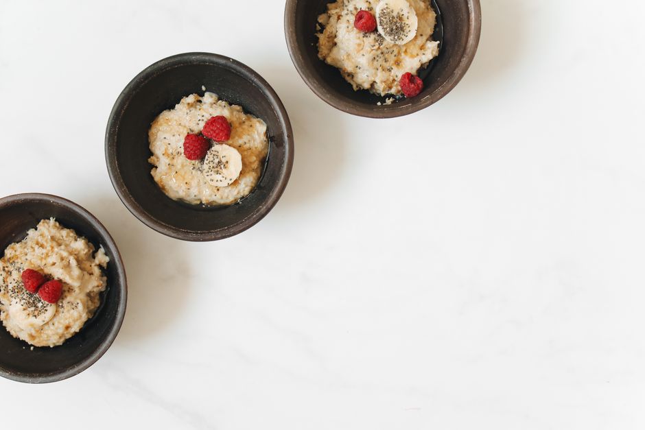 Três porções de mingau decoradas com frutas vermelhas e fatias de banana em tigelas de cerâmica, apresentadas sobre uma superfície branca.