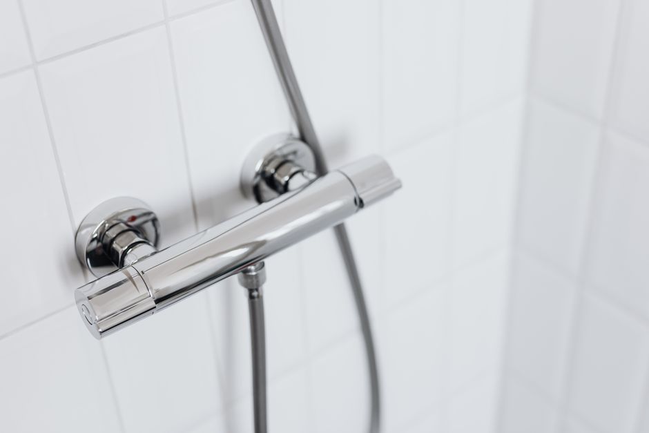 Foto de uma ducha de banheiro com acabamento cromado instalada em uma parede de azulejos brancos, representando higiene e climatização de ambientes sanitários.