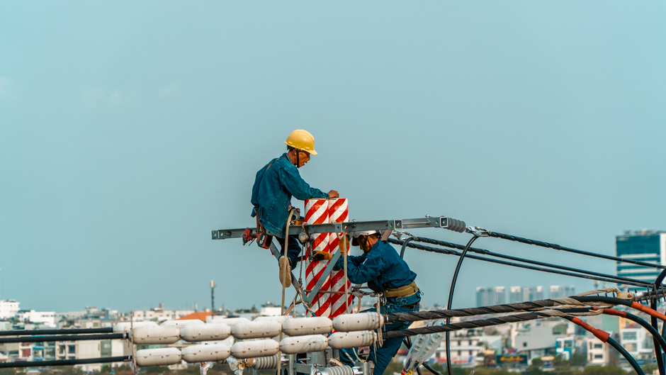 Imagem de operários trabalhando na instalação de infraestrutura elétrica em um canteiro de obras com vista urbana ao fundo.