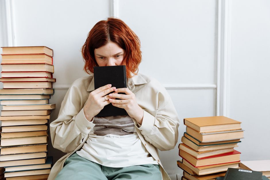 Mulher com cabelo cacheado e ruivo lendo um livro, cercada por pilhas de outros livros, em um ambiente de estudo ou pesquisa.