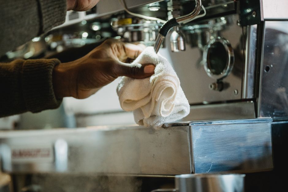 Pessoa limpando uma máquina de café espresso com um pano de prato, destacando a manutenção e higiene em estabelecimentos gastronômicos.