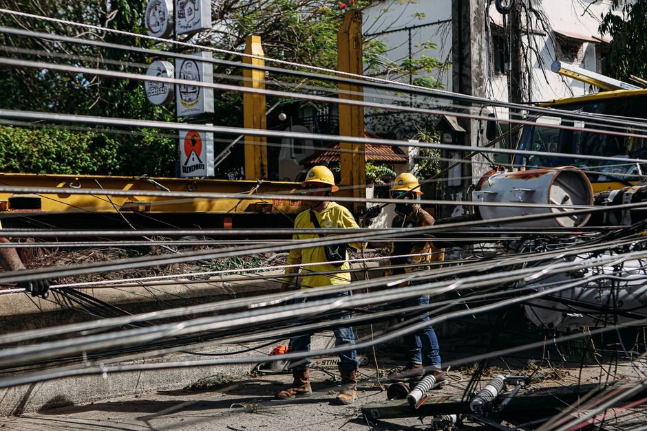 Imagem de trabalhadores em manutenção elétrica em uma área urbana, destacando a intervenção em cabos de energia e infraestrutura de rede elétrica.
