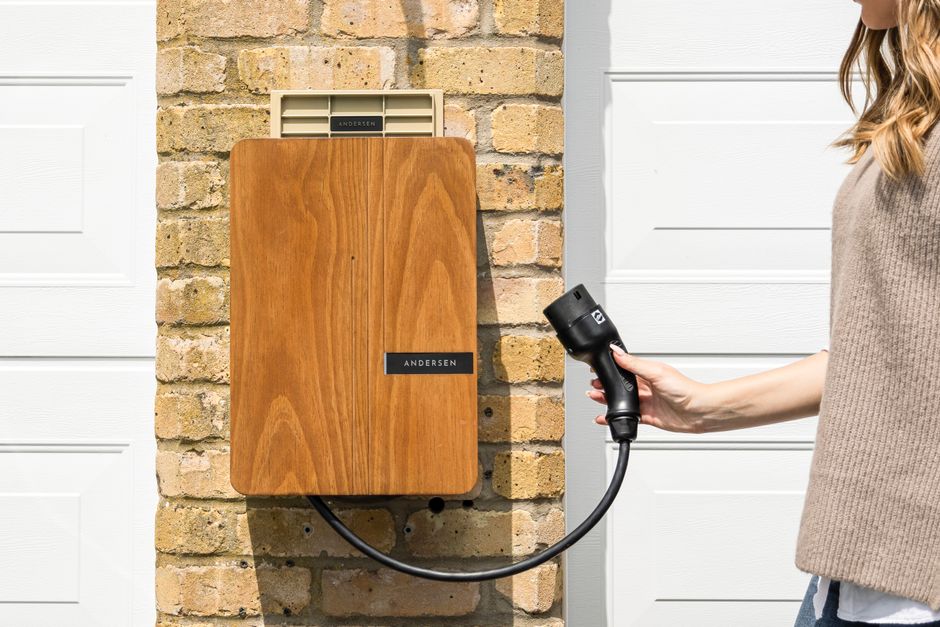 Imagem de uma pessoa carregando um carregador de carro elétrico próximo a uma parede de tijolos e uma porta de garagem branca, simbolizando sustentabilidade e mobilidade elétrica.