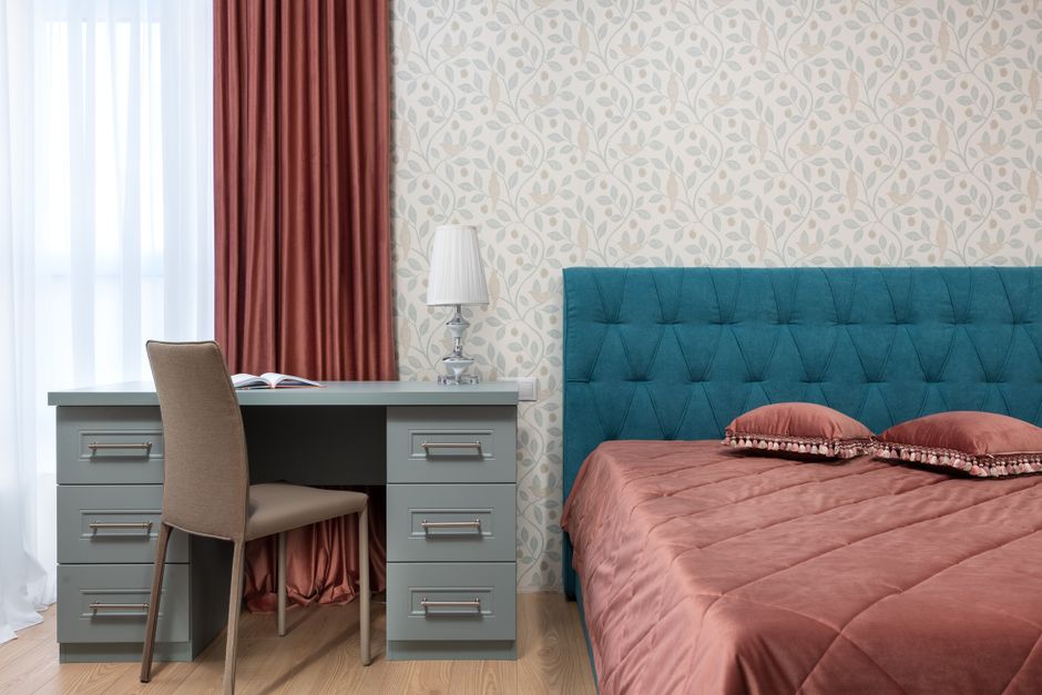 Quarto aconchegante com parede de papel de parede com padrão vegetal, mesa de trabalho com luminária e quarto com cabeceira azul e edredom cor de rosa.
