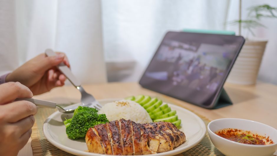 Pessoa jantando uma refeição equilibrada enquanto assiste a uma apresentação de slides ou vídeo em um tablet, em ambiente interno com iluminação natural.