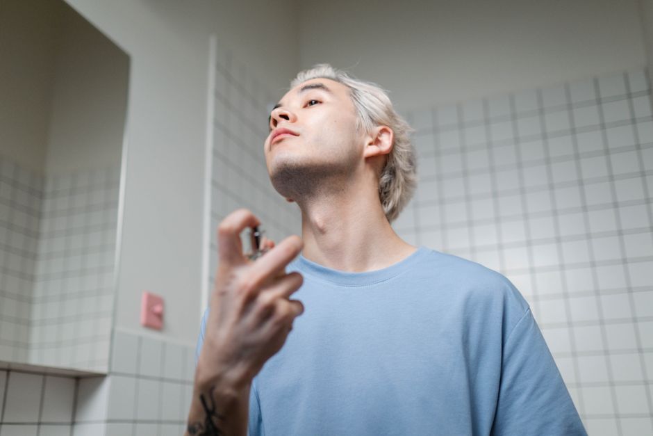 Homem utilizando um perfurador de fragrância ou spray de perfume no rosto, em um ambiente com azulejos brancos, sugerindo uma rotina de autocuidado ou rotina de beleza.