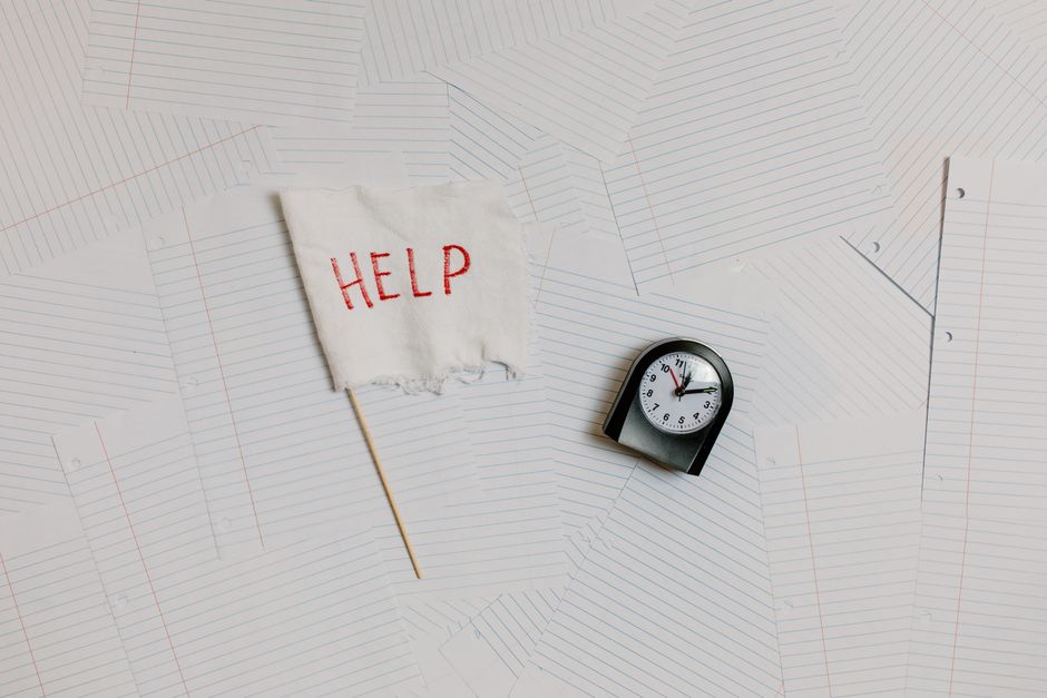 Imagem com um cartaz escrito 'HELP' e um temporizador sobre uma superfície repleta de papéis com linhas, sugerindo urgência ou crise de tempo.
