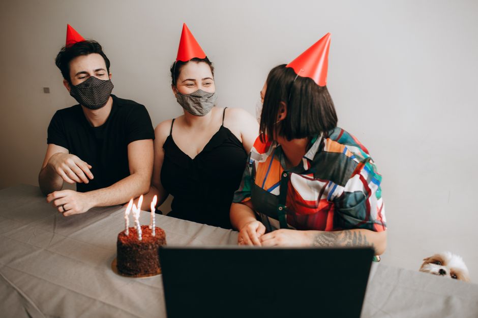 Grupo de amigos comemorando aniversário virtualmente com festa e velas, usando chapéus de festa e máscaras faciais.