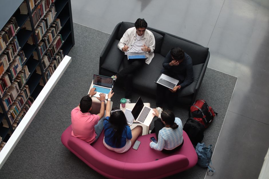 Grupo de estudantes ou profissionais realizando estudo ou trabalho em um espaço de biblioteca ou lounge acadêmico.