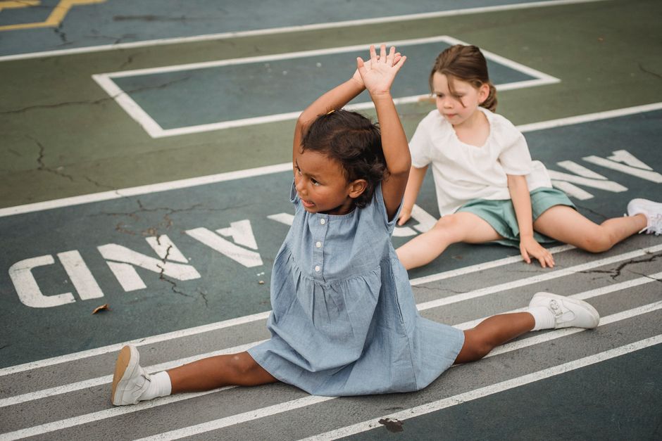 Duas crianças em uma brincadeira de alongamento no chão de um parque de diversões com marcações de pista de corrida, demonstrando brincadeira e diversão infantil.
