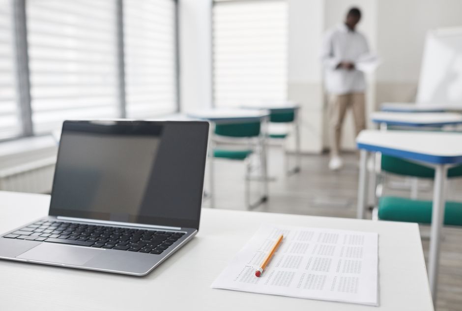Ambiente de sala de aula moderna com foco em estudos e preparação acadêmica, com destaque para um notebook, documentos e um estudante ao fundo.