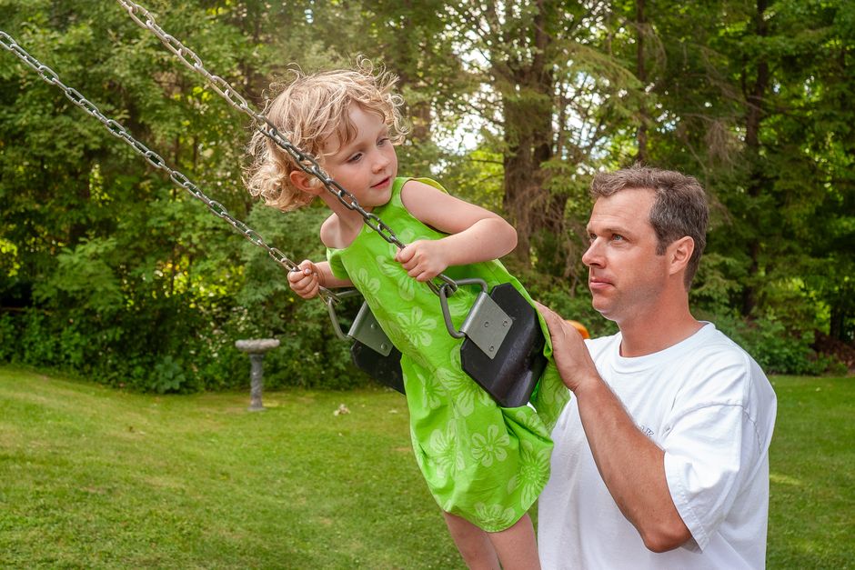 Pai brincando com a filha no balanço ao ar livre em um ambiente com árvores verdes.