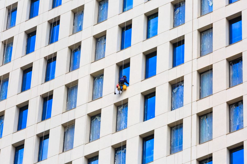 Profissional de limpeza realizando conservação de janelas em edifício corporativo alto, simbolizando manutenção e cuidados com infraestrutura urbana.