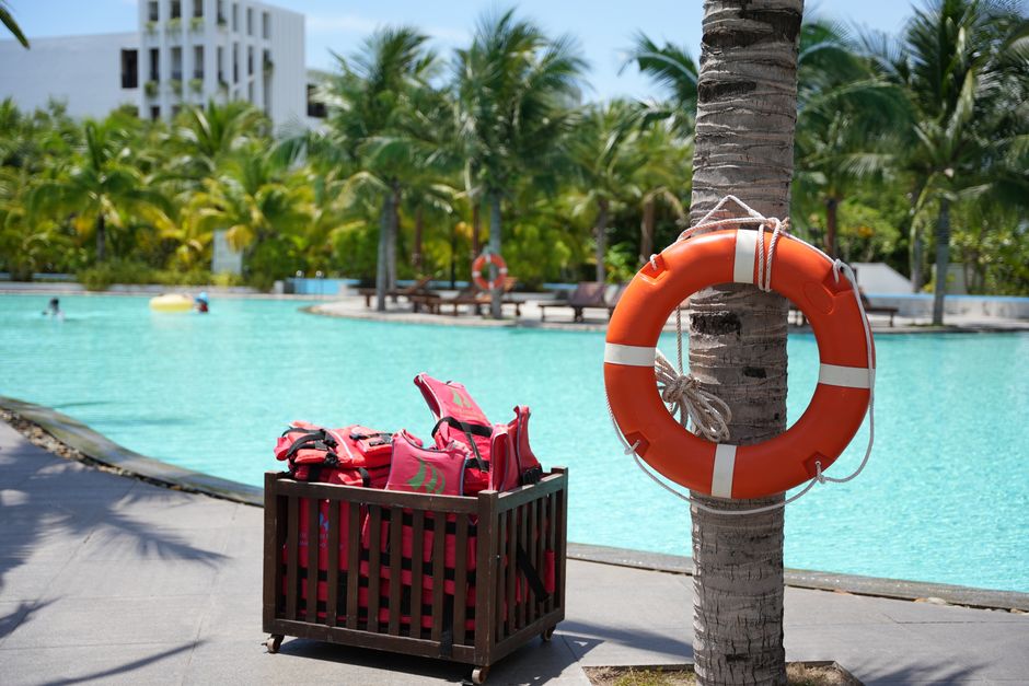 Imagem de uma área de lazer na piscina de um resort, com equipamentos de salva-vidas e um cenário tropical ao fundo.