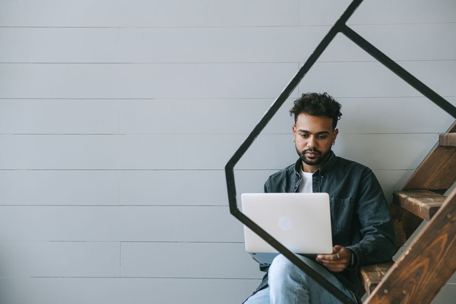 Jovem profissional estudando ou trabalhando com laptop em um ambiente moderno e minimalista, próximo às escadas de madeira.