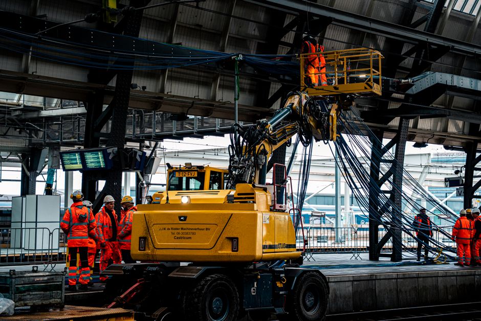 Imagem de uma equipe de trabalhadores usando equipamentos de proteção em uma construção ou manutenção de infraestrutura industrial, com uma máquina de elevação no ambiente fechado.