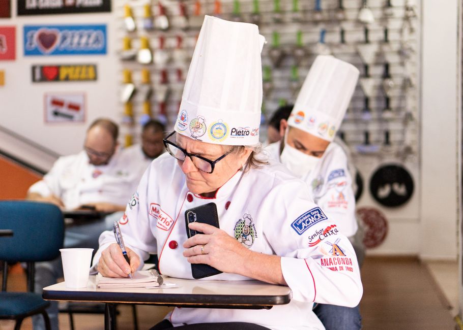 Chef profissional observando o celular enquanto anota informações, em ambiente de cozinha de confeitaria ou padaria.