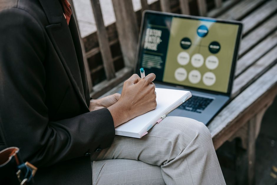 Pessoa analisando dados de planejamento financeiro ao ar livre, com laptop exibindo gráficos e anotações em um caderno.