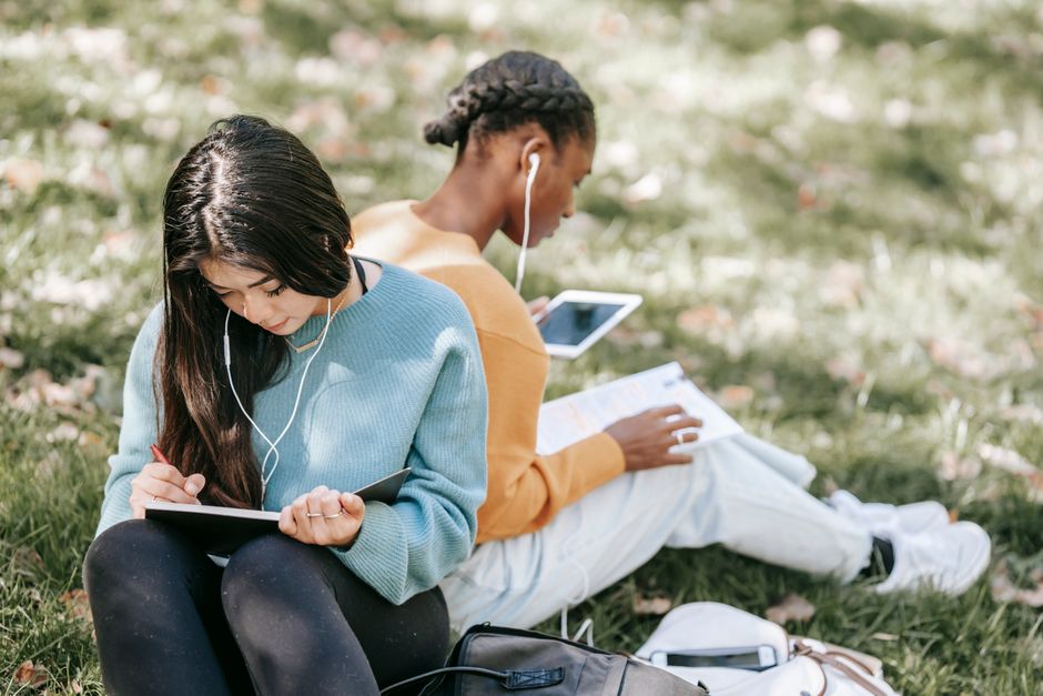 Dois jovens sentados ao ar livre, cada um usando dispositivos eletrônicos, aprendendo ou estudando de forma colaborativa e descontraída.