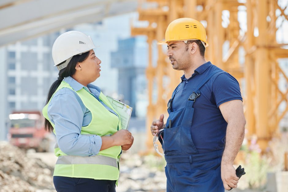 Duplo de engenheiros discutindo em um canteiro de obras com estrutura de madeira ao fundo, representando temas de construção civil e gestão de obras.