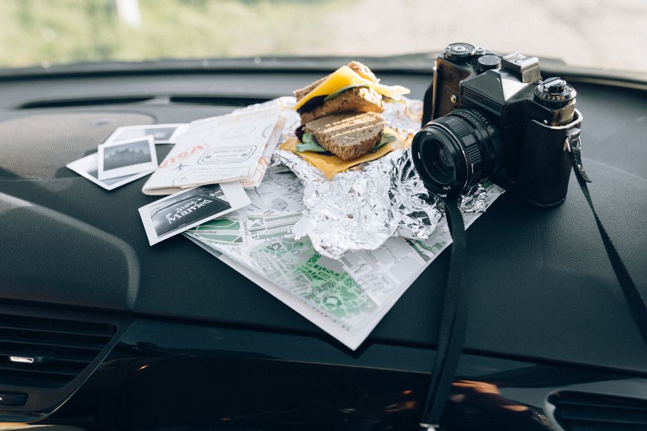 Foto de um dashboard de carro com um lanche, câmera fotográfica, mapas, fotos de viagem, e documentos, sugerindo um momento de viagem ou aventura.